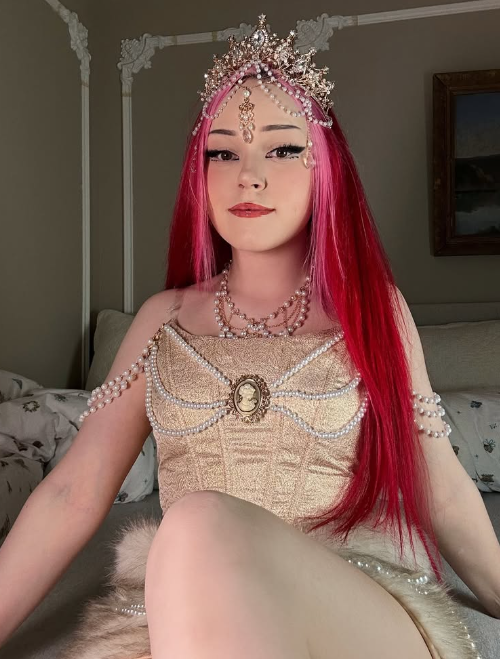 Alice Starr tar et bilde mens hun sitter på sengen sin i et beige vintage cosplay-antrekk og en perle på hodet.