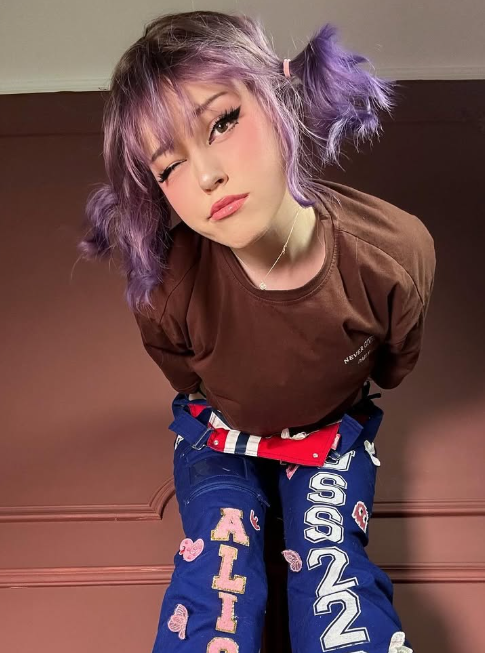Alice Starr tar et bilde i en brun topp og blå bukser og har på seg en lilla kort parykk.
