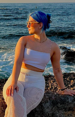 @nooojia tar et bilde pÄ stranden, sittende pÄ en stein og kledd i en hvit crop-topp og hvite strandbukser.