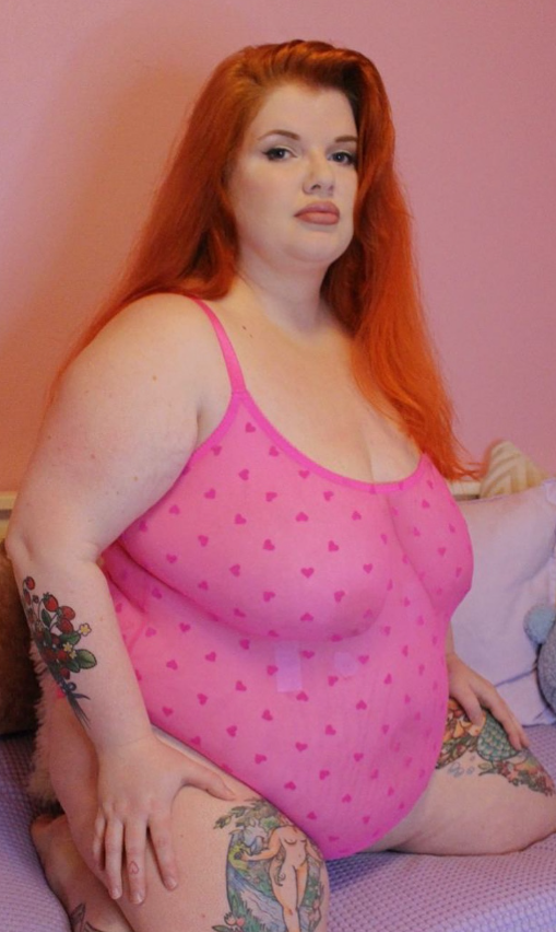 Ginger Minx tar et bilde mens hun sitter på sengen sin og er kledd i et sexy rosa antrekk.
