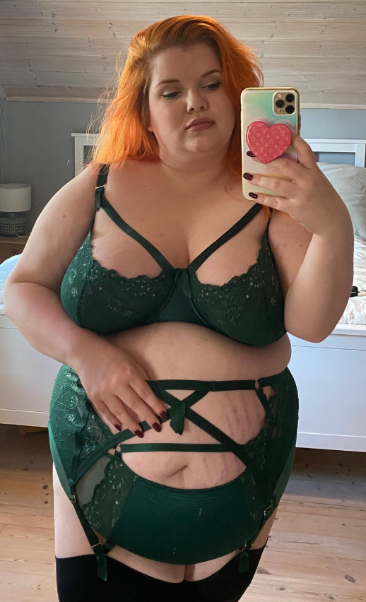 Ginger Minx tar et bilde mens hun står foran sengen sin i sexy grønt undertøy mens hun holder telefonen i hånden.